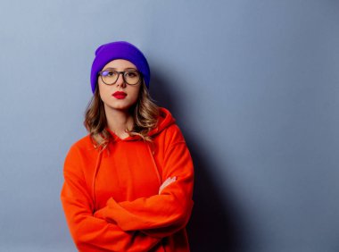 Turuncu kapüşonlu stil kızı ve gri arka planda mor şapkalı.