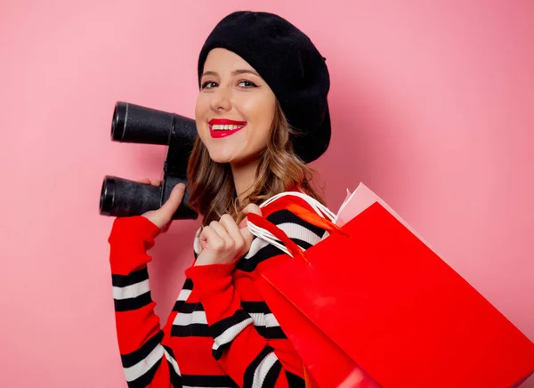 Mujer joven con prismáticos y bolsas de compras sobre fondo rosa —  Fotos de Stock