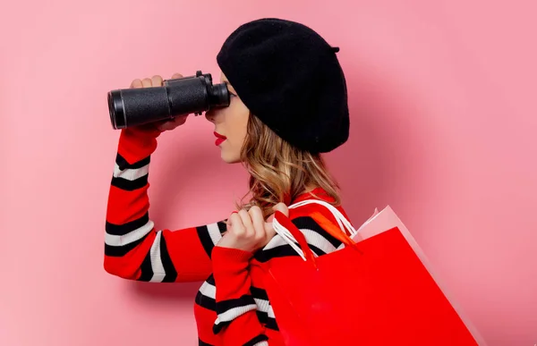 Jovem com binóculos e sacos de compras em fundo rosa — Fotografia de Stock