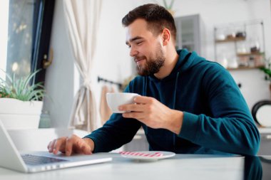 Genç adam mutfağındaki bilgisayarda çalışıyor ve çay içiyor.