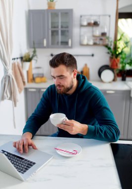 Genç adam mutfağındaki bilgisayarda çalışıyor ve çay içiyor.