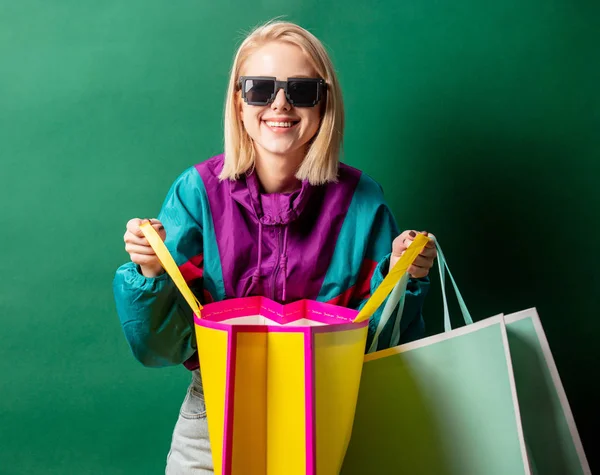 Blond meisje in jaren 90 stijl jas met boodschappentassen op groene rug — Stockfoto