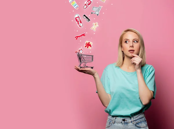 Mooie blonde vrouw verzamelen geschenken in een kar op roze backgroun — Stockfoto