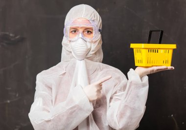 Koruyucu giysili ve maskeli bir kadın alışveriş sepeti tutuyor. 