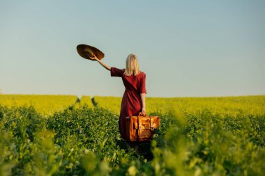 Kırmızı elbiseli, şapkalı, kolza tohumu tarlasında bavullu sarışın bir kadın. 