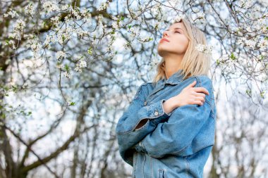 Baharda çiçek açan ağacın yanında kot elbiseli sarışın kadın. 