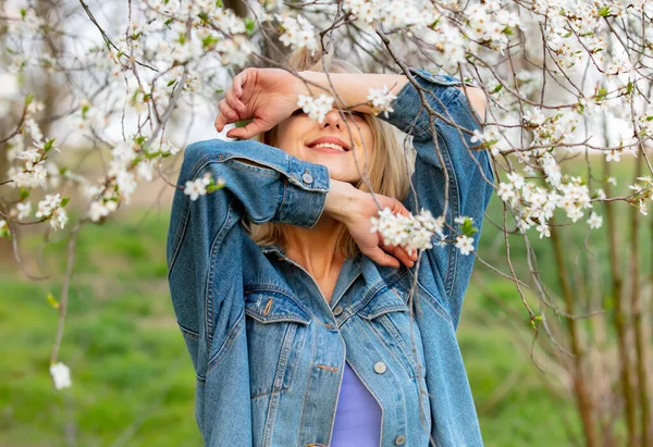 春の時間に咲く木の横にデニム服のブロンドの女性 — ストック写真
