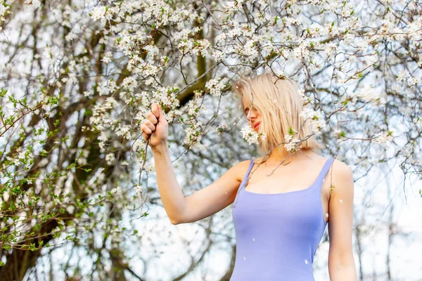 春に咲く木の横に青いドレスの美しいブロンドの女性 — ストック写真
