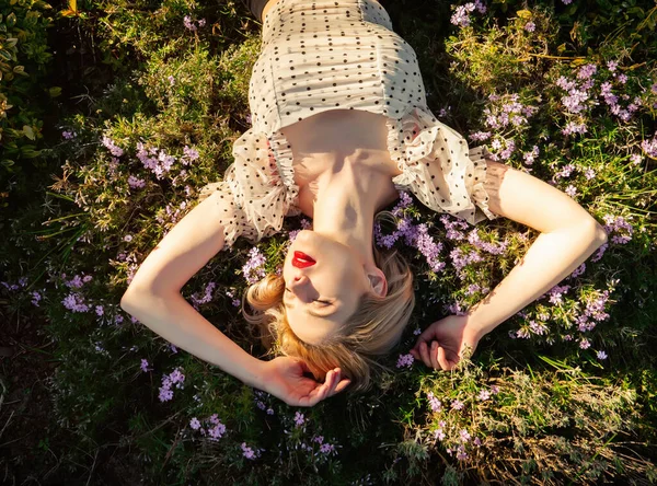 ブロンドの女の子は日没の田舎で花と草の上に横たわっています — ストック写真