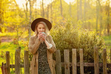 Günbatımında bahçede bir fincan kahveyle şapkalı ve ceketli şık sarışın kadın.
