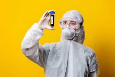 Koruyucu giysi ve maske giyen doktor sarı arka planda kanla dolu kavanozu tutuyor.
