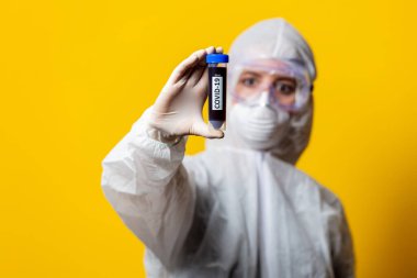 Koruyucu giysi ve maske giyen doktor sarı arka planda kanla dolu kavanozu tutuyor.