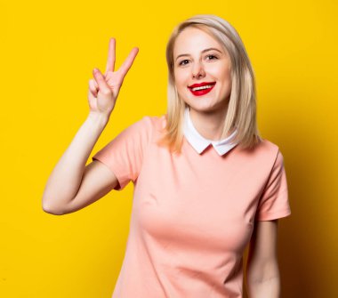 Pembe elbiseli sarışın kız sarı arka planda jest yapıyor. 