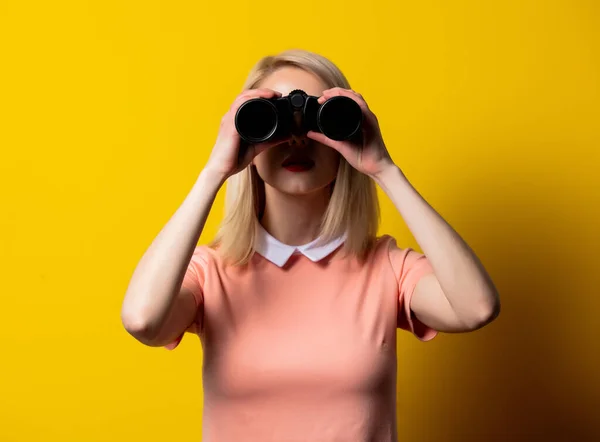 穿着粉色衣服的金发女孩穿着黄色背景的双筒望远镜注视着 — 图库照片