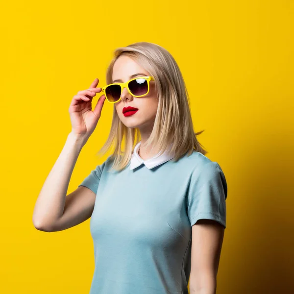 ブロンド女の子でサングラスと青いドレス上の黄色の背景 — ストック写真