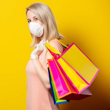 Sarı arka planda alışveriş torbaları olan maskeli sarışın kız..