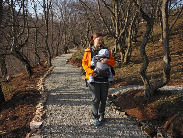 Mother with baby in baby carrier walks by the forest road — Stock Photo, Image