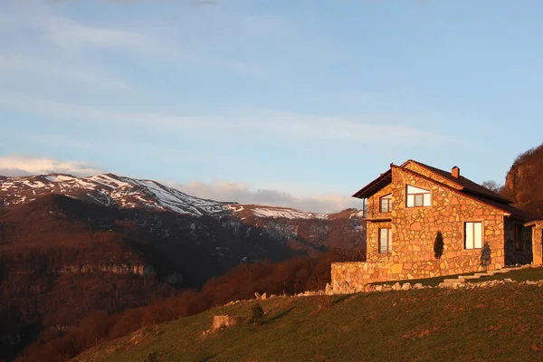 Solitaria casa de piedra en la colina en el fondo de las montañas nevadas, al amanecer, horizontal — Foto de Stock