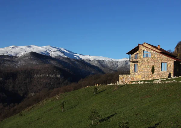 Solitaria casa de piedra en la colina en el fondo de las montañas nevadas, cielo azul claro, horizontal — Foto de Stock