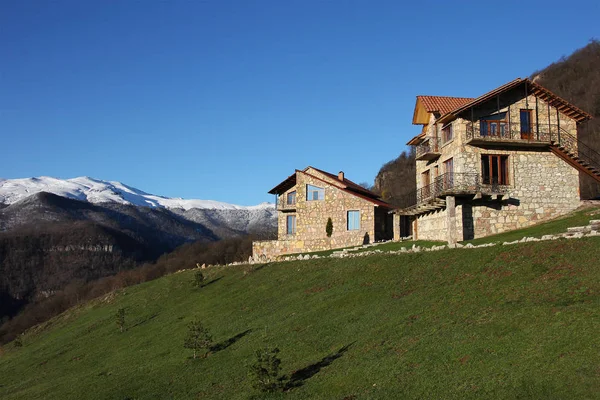 Dos casas de piedra en la colina en el fondo de las montañas nevadas, cielo azul claro, horizontal — Foto de Stock