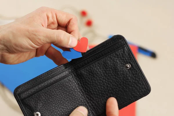 Menselijke hand houdt twee papieren harten rood en blauw, probeert om het in zwart lederen portemonnee, concept valentijn cadeau of een goede investering — Stockfoto
