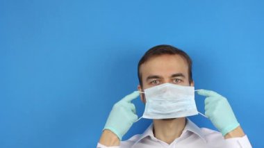 Erkek yüzüne ön görüş, tıbbi maske takar ve ameliyat eldivenlerini takar, doktor virüs koronavirüsüne veya kirliliğe karşı korunduğunu gösterir, başlıklar için mavi arkaplan