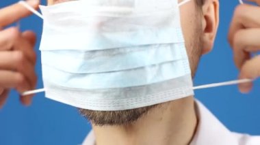 Erkek yüzünün yakın plan görüntüsü, tıbbi maskeyi nasıl taktığını, virüsü ya da kirliliği nasıl önlediğini ya da koruduğunu, mavi arka plan 