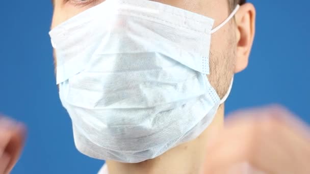 男性面部特写部分 演示如何去除使用过的医疗面罩 卫生规则以预防或保护病毒或污染 蓝色背景 — 图库视频影像