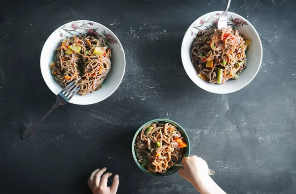 simple family dinner or lunch, buckwheat vegan noodles with vegetables and beans, three plates and forks, child hands, black background, top view above