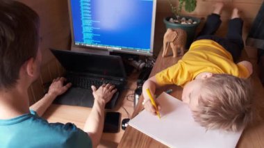 Baba evde bilgisayarda çalışıyor, küçük çocuk yanında uzanıyor ve albümde resim yapıyor, salgın ya da karantinada evde kalma konsepti, kendini soyutlamış aile içi hayat tarzı, ebeveynlik.