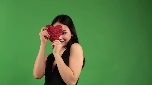 Menina asiática segurando um coração vermelho em suas mãos cobrindo-o com um olho e sorrindo, conceito de celebração da sexualidade — Fotografia de Stock