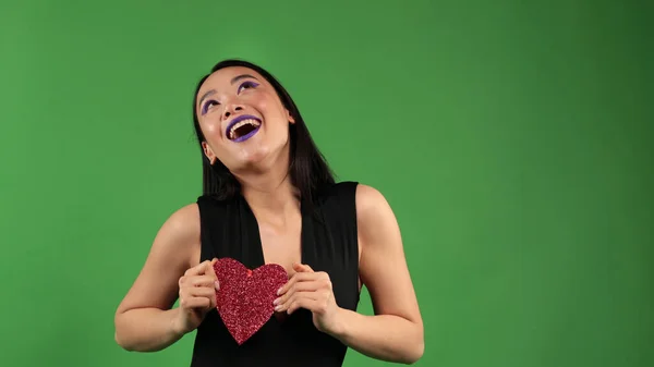 Asiático sorrindo mulher dançando com um papel vermelho coração em suas mãos e cobrindo seu rosto com ele em um fundo verde isolado, conceito do feriado dos amantes — Fotografia de Stock