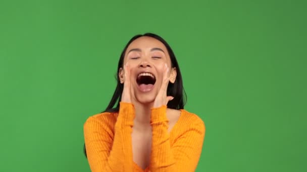 Young Asian woman screams in surprise and fun, substituting both hands to her mouth, isolated on a green background — Wideo stockowe