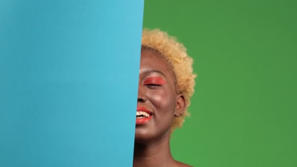 Zbliżenie twarzy ciemnoskórej młodej kobiety uśmiechniętej i w połowie pokrytej niebieskim tłem na odizolowanym zielonym tle — Wideo stockowe