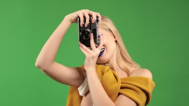 O tânără blondă cu o cameră în mâini face o fotografie și zâmbește la camera foto pe un fundal verde izolat, conceptul unui călător — Videoclip de stoc