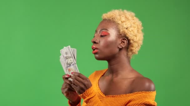 Mujer afroamericana con un fan del dinero ondea dinero y lo dispersa con una expresión complacida en un fondo verde aislado, concepto de riqueza y ganar — Vídeos de Stock