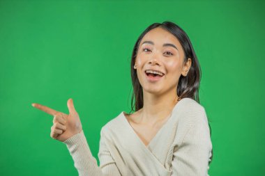 Beyazlar içindeki gülümseyen ve kendine güvenen kadın kameraya bakan ürününüzü işaret ediyor. Yeşil arka planda izole edilmiş. Hoş karışık Asyalı, Avrupa modeli