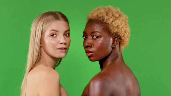 Närbild porträtt av två vackra unga blonda och mörkhyade kvinnor, kvinnor som står med varandra halv vända med nakna axlar och ler mot kameran på isolerad grön bakgrund — Stockfoto
