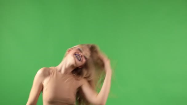 Красивая молодая женщина в стильной одежде качает головой с длинными светлыми волосами и танцует улыбаясь в камеру и изолированный зеленый фон — стоковое видео