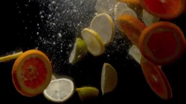 Dilimlenmiş taze limon ve portakallar siyah arka planda suya düşüyorlar.