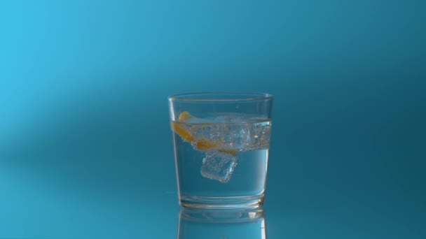 氷とレモンのスライスを青い背景にミネラル輝く水でガラスの中に回転させます 天然炭酸ミネラルウォーター — ストック動画