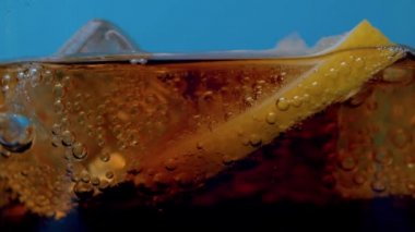 Bardakta yükselen gaz baloncukları ve kola çok yakından geliyor. Dilimlenmiş limon ve buz parçaları mavi arka planda kokain yüzeyinde yüzüyor. Gazlı içecek, bardağa doldurulmuş.