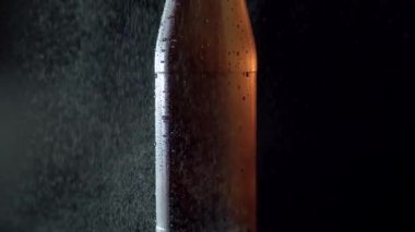 Close up misted glass bottle spraying with water on a black background. Drops of water falling down and beautifully shining in a ray of light. Craft beer closeup