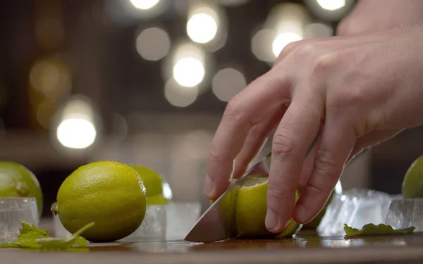 Bulanık ampullerin arka planında erkek el limonu dilimleme masasını kapatın. Tazeleyici içecekler pişiriyorum. Barmen yakın planda çalışıyor.