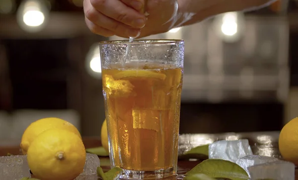 Erkek elinin limonu buzlu çay ya da kokteyl ile bardağa sıkıştırmasını kapat. Tazeleyici bir içecek, buz, limon ve yapraklarla dolu bir bardak bulanık ışık arka planında.