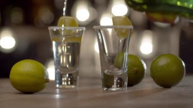 Barmene yakın bardaklara tekila dolduruyor ve güçlü bir alkollü içeceği ateşe veriyor. Bulanık ampullerin arkaplanındaki çubuk sayacı.
