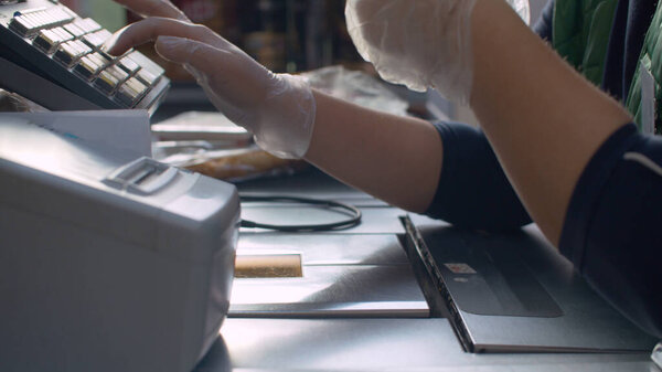 Close up checkout counter in the supermarket during coronavirus epidemic. Cashier's hands in protective gloves working on cash register. COVID-19 pandemic