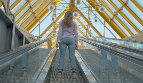Молодая женщина стоит на движущемся эскалаторе. Вид сзади, за выстрелом. Блондинка с длинными волосами в современном здании
