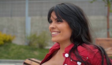 Açık hava bankında oturan güzel esmer, gülümseyen kadın portresi. Şehirdeki gerçek insanlar..