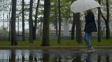 Ellerinde şemsiyesi olan neşeli bir kadın su birikintisinde yürüyor ve ayaklarıyla su fışkırtıyor. Parkta yağmurlu bir gün. Gerçek insanlar.